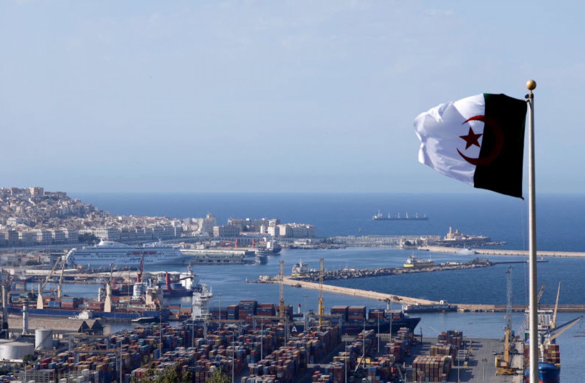 The national flag of Algeria flies near the Bay of Algiers against the backdrop of the old town known as the Casbah and cargo containers in Algiers, Algeria, on Aug. 25, 2022.