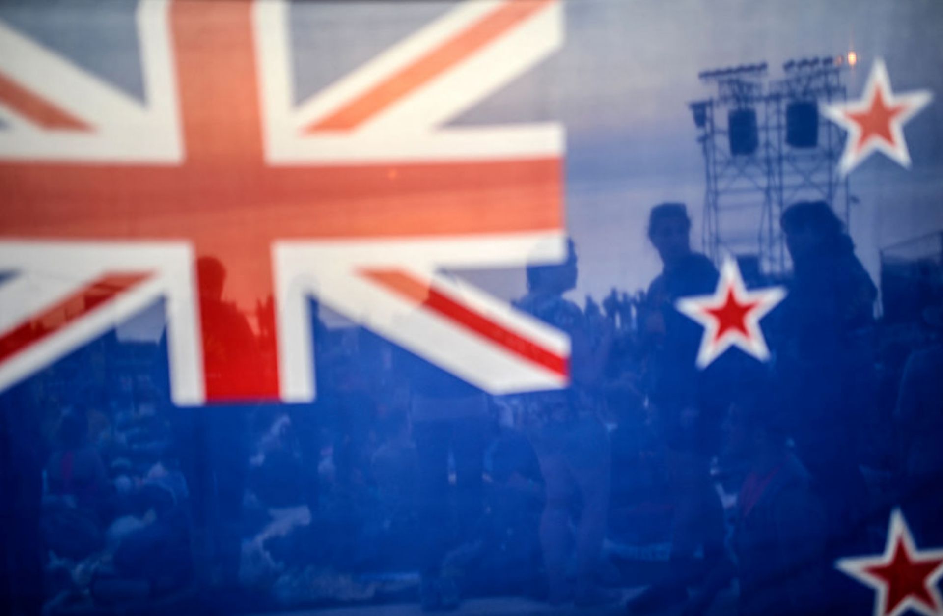 People are seen behind a New Zealand flag during a ceremony celebrating the anniversary of Anzac Day. People are seen behind a New Zealand flag during a ceremony celebrating the anniversary of Anzac Day.