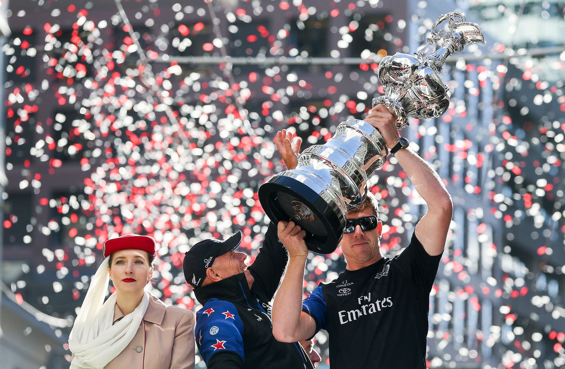 Peter Burling, the helmsman of Emirates Team New Zealand, flanked by team CEO Grant Dalton (r) and a representative from sponsor Emirates airlines, holds aloft the America's Cup during a victory parade in Wellington, New Zealand. 