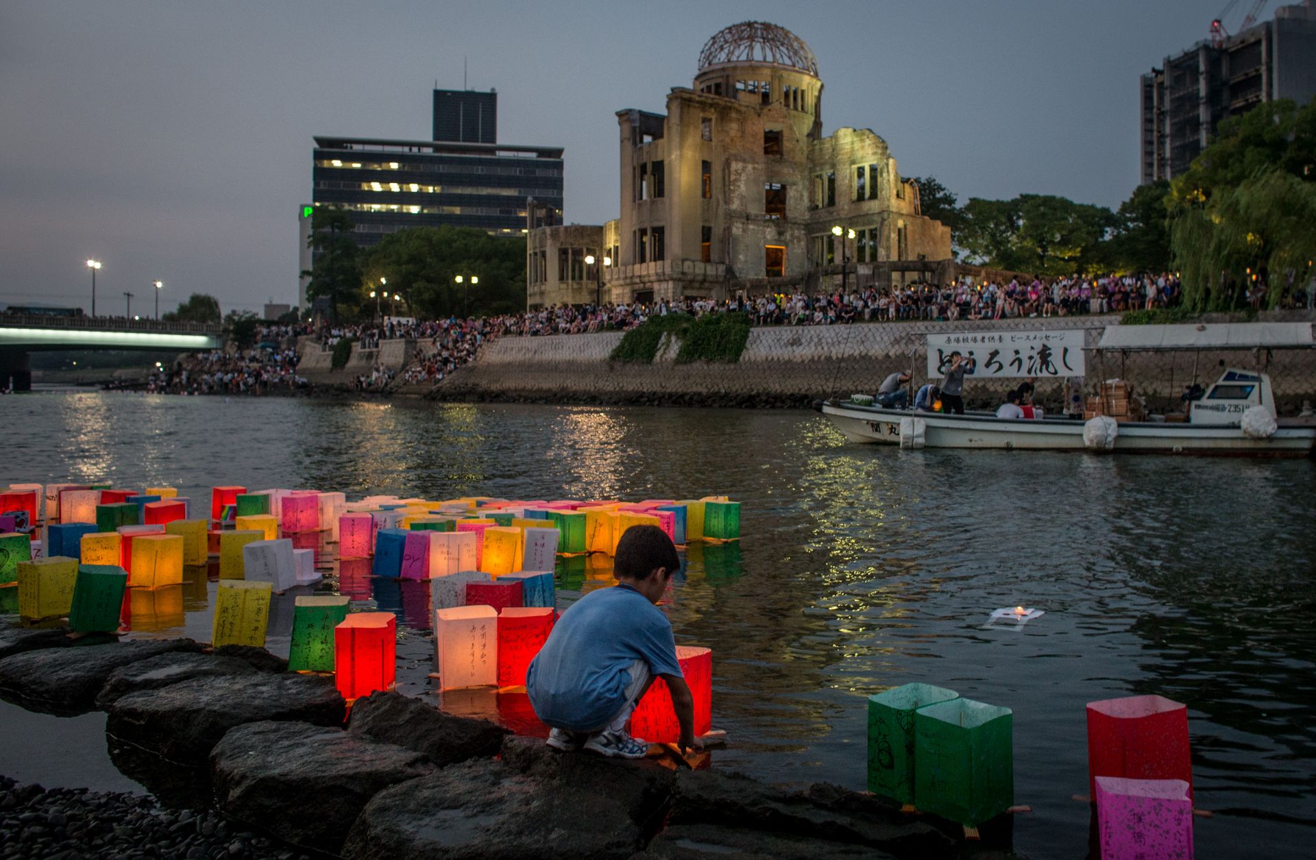 A boy floats a candle lit paper lantern on the river in front of the Atomic Bomb Dome.