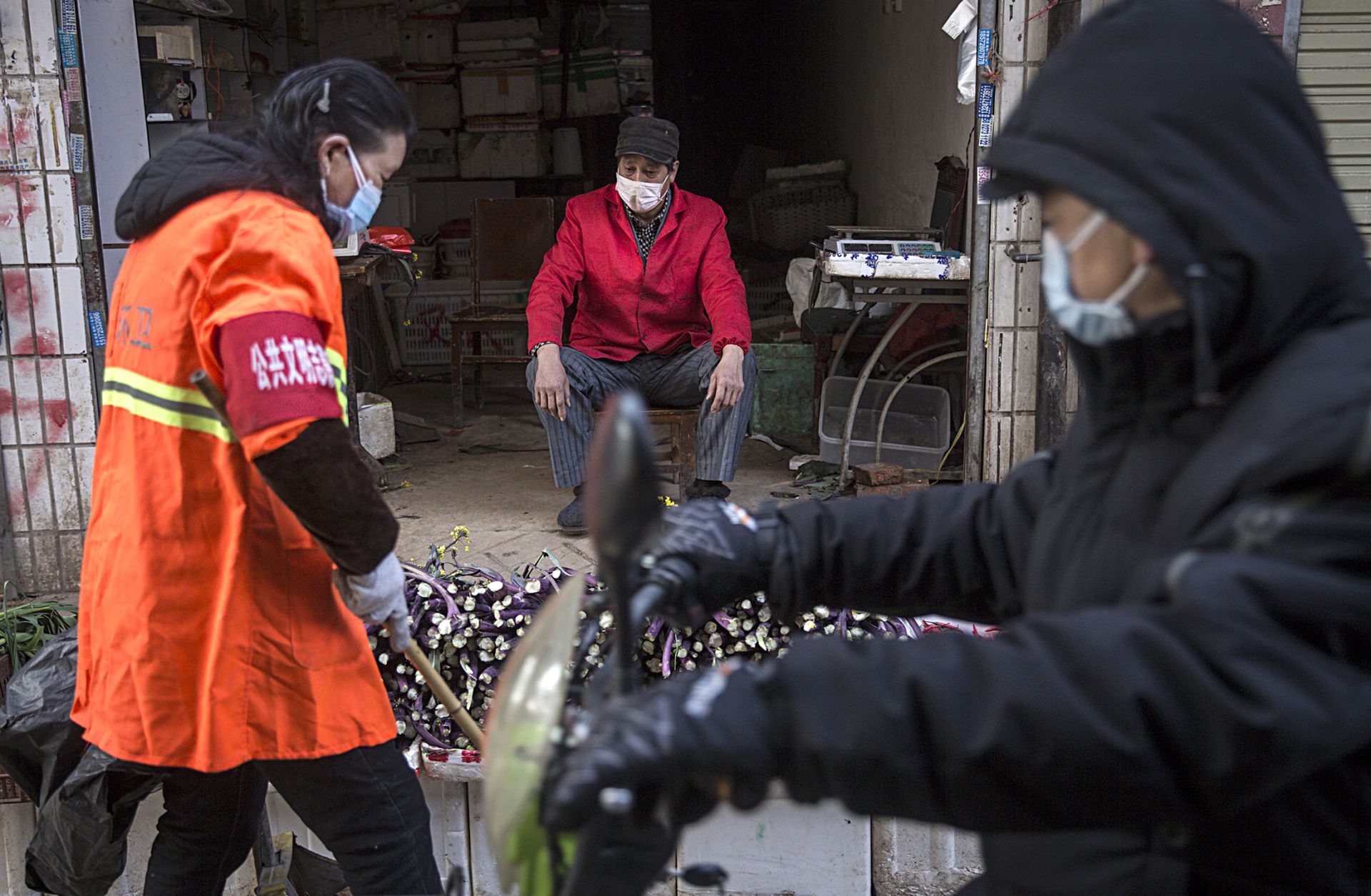 A customer examines a produce vendor's wares at a market in Wuhan, China. The economic effects of the city's quarantine on the country's larger economy will grow more pronounced the longer the lockdown stays in place. This photo shows a masked vendor and customers of his wares in an alley in Wuhan, China, on January 31, 2020.