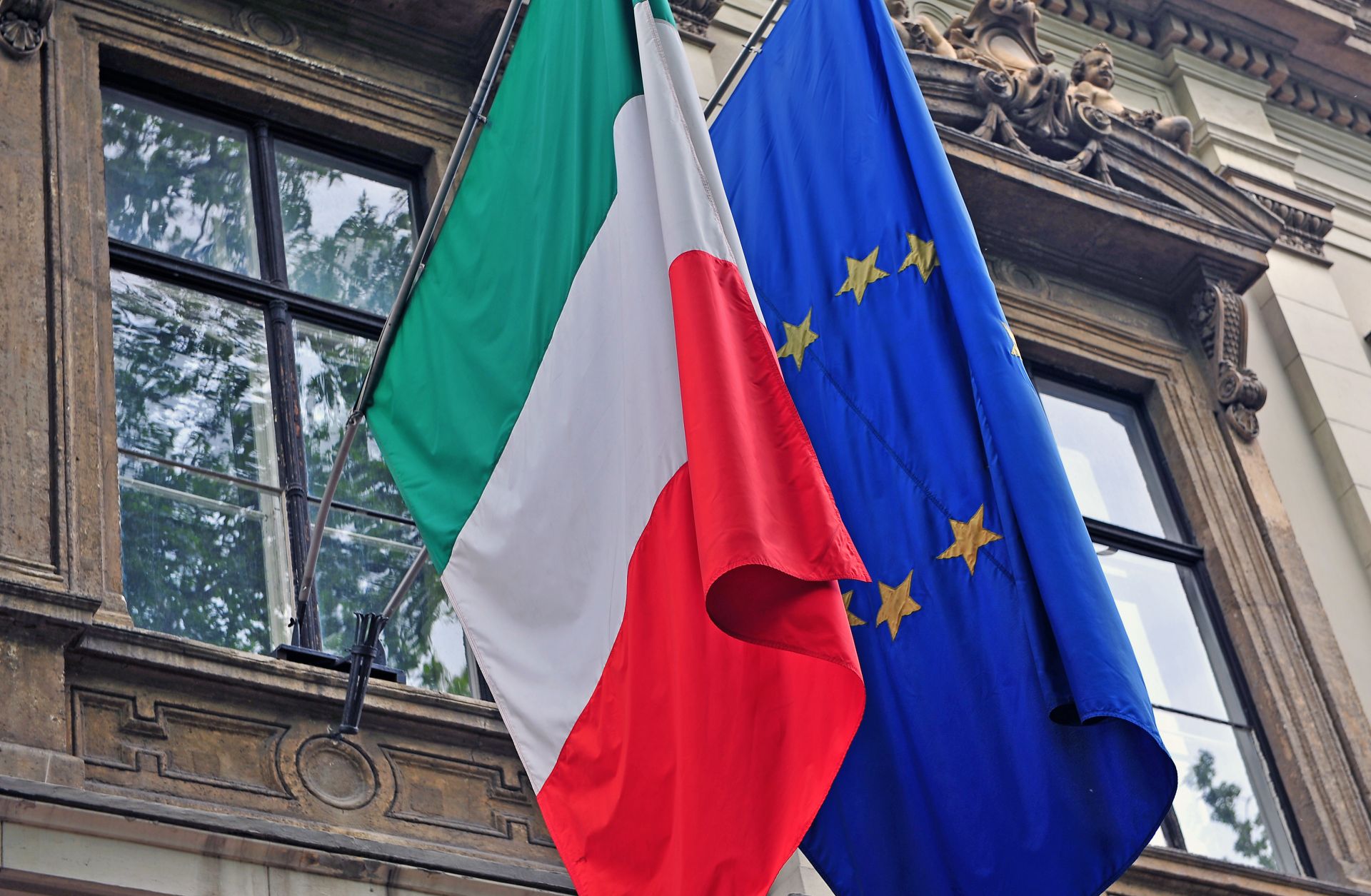 The Italian and EU flags hang side by side from the facade of a government building in Italy.