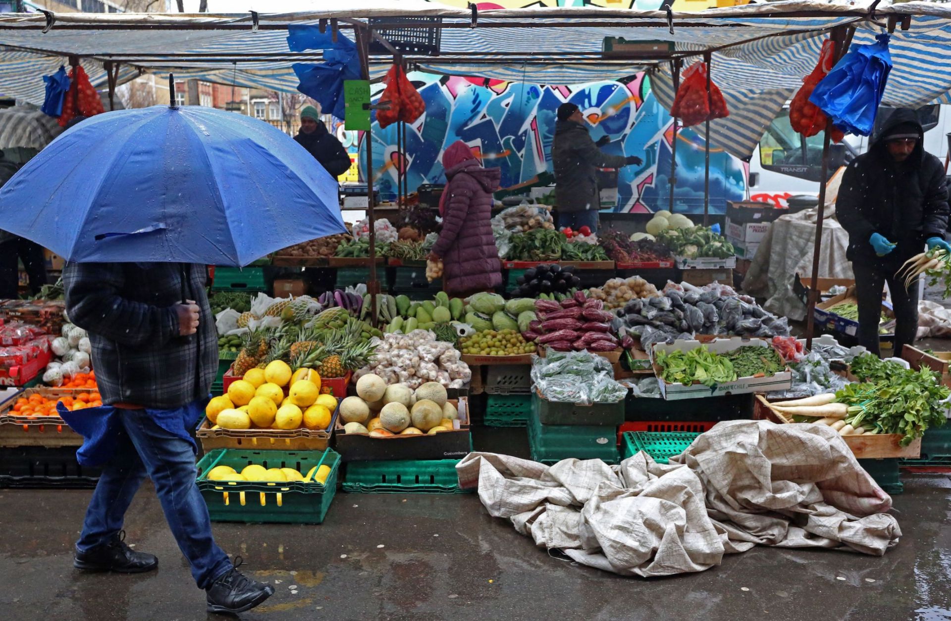 A pedestrian shelters from the rain as they walk past fruit and vegetables displayed for sale at a market in east London on March 31, 2023.