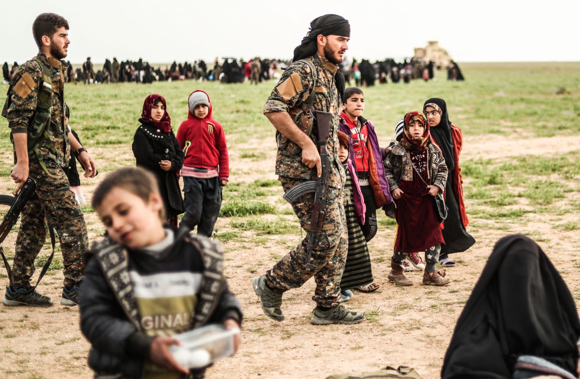 Fighters with the Syrian Democratic Forces walk past civilians at a screening area for evacuees from the Islamic State's embattled holdout in Baghouz, Syria, on Feb. 26, 2019. 