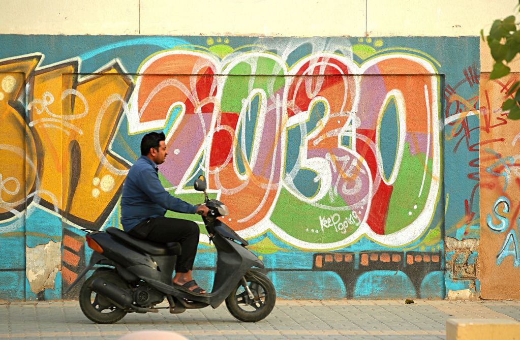 This photo shows a scooter rider passing graffiti alluding to the Saudi Vision 2030 modernization program