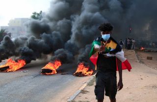 A protester draped with Sudan’s national flag stands in front of burning tires during a demonstration in Khartoum on Oct. 25, 2021. A protester draped with Sudan’s national flag stands in front of burning tires during a demonstration in Khartoum on Oct. 25, 2021.