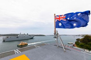 An Australian flag waves as a warship transits a waterway in the background in a photo taken on Jan. 18, 2020. 