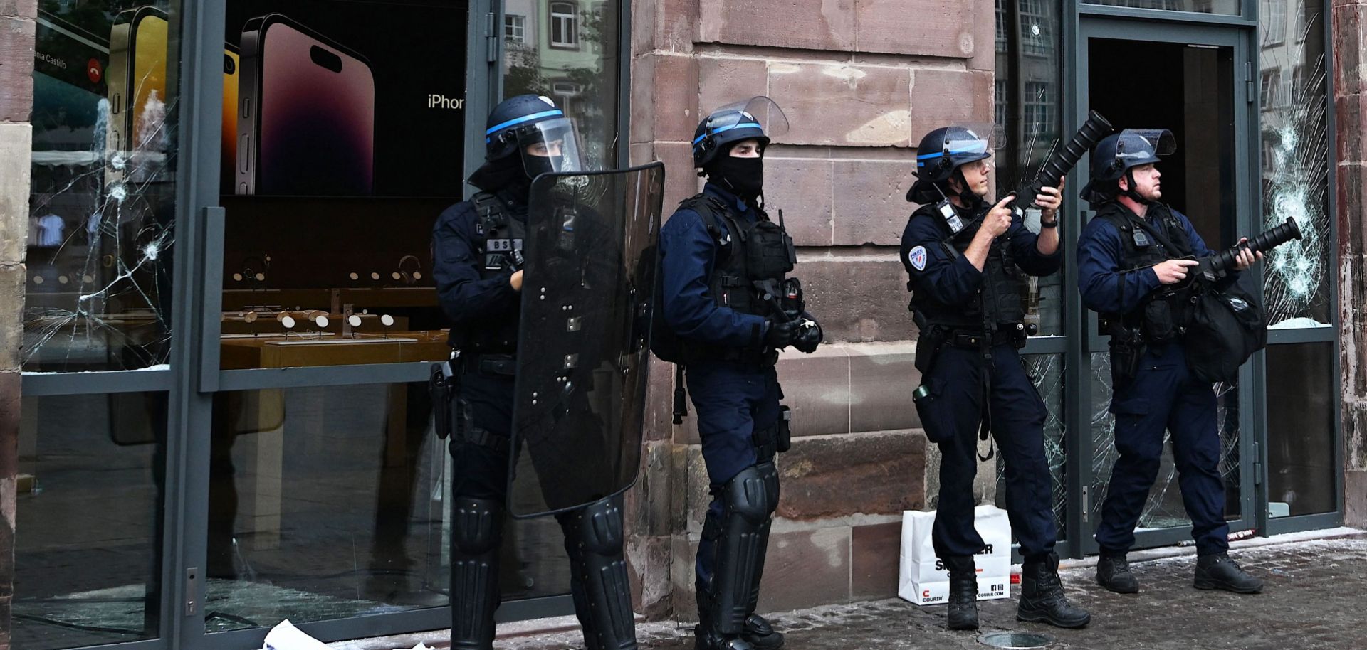 French police officers in riot gear stand guard and hold tear gas canister launchers next to the facade of a damaged Apple Store in Strasbourg, eastern France, on June 30, 2023.