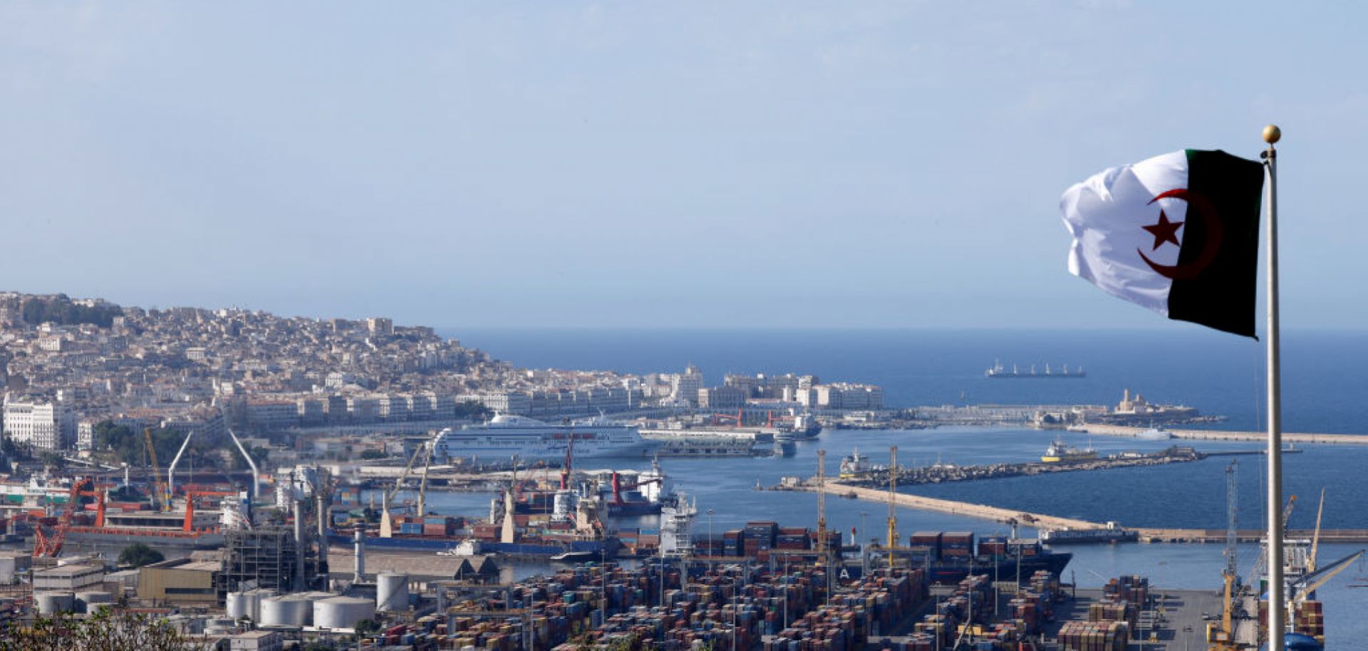 The national flag of Algeria flies near the Bay of Algiers against the backdrop of the old town known as the Casbah and cargo containers in Algiers, Algeria, on Aug. 25, 2022.