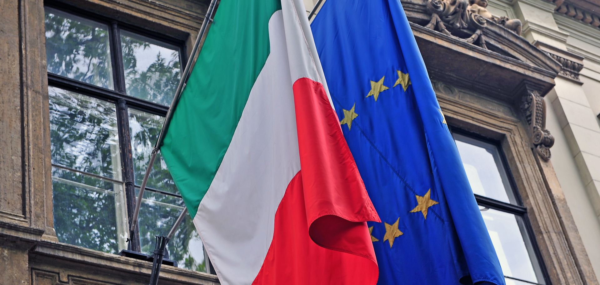 The Italian and EU flags hang side by side from the facade of a government building in Italy.