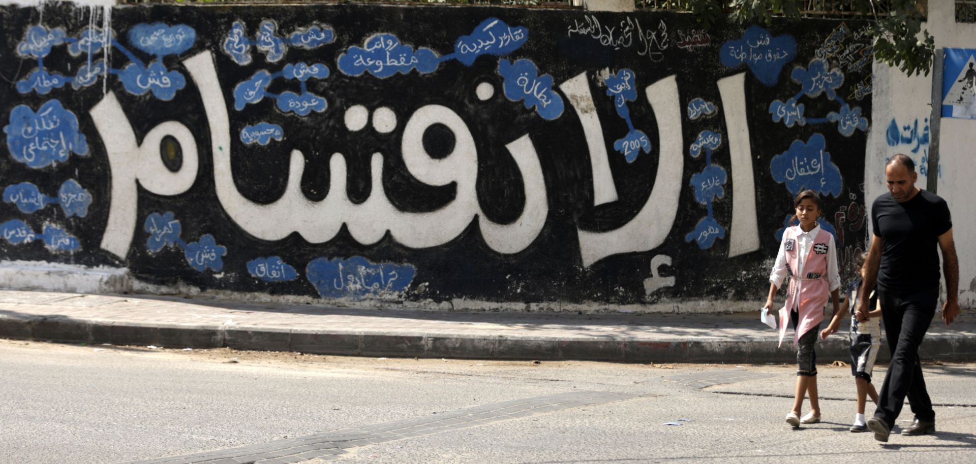 A Palestinian man walks through Gaza with two of his daughters in front of a wall graffitied with the Arabic word for division.