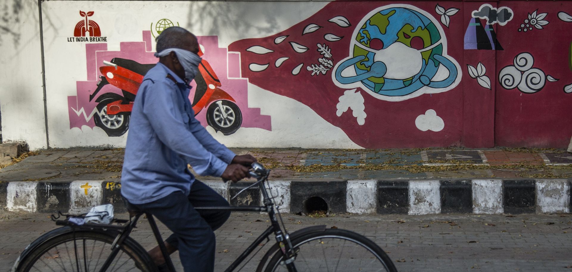 A man wearing a bandana peddles his bicycle in front of a mural depicting the globe covered in a mask on April 13, 2020, in New Delhi, India.