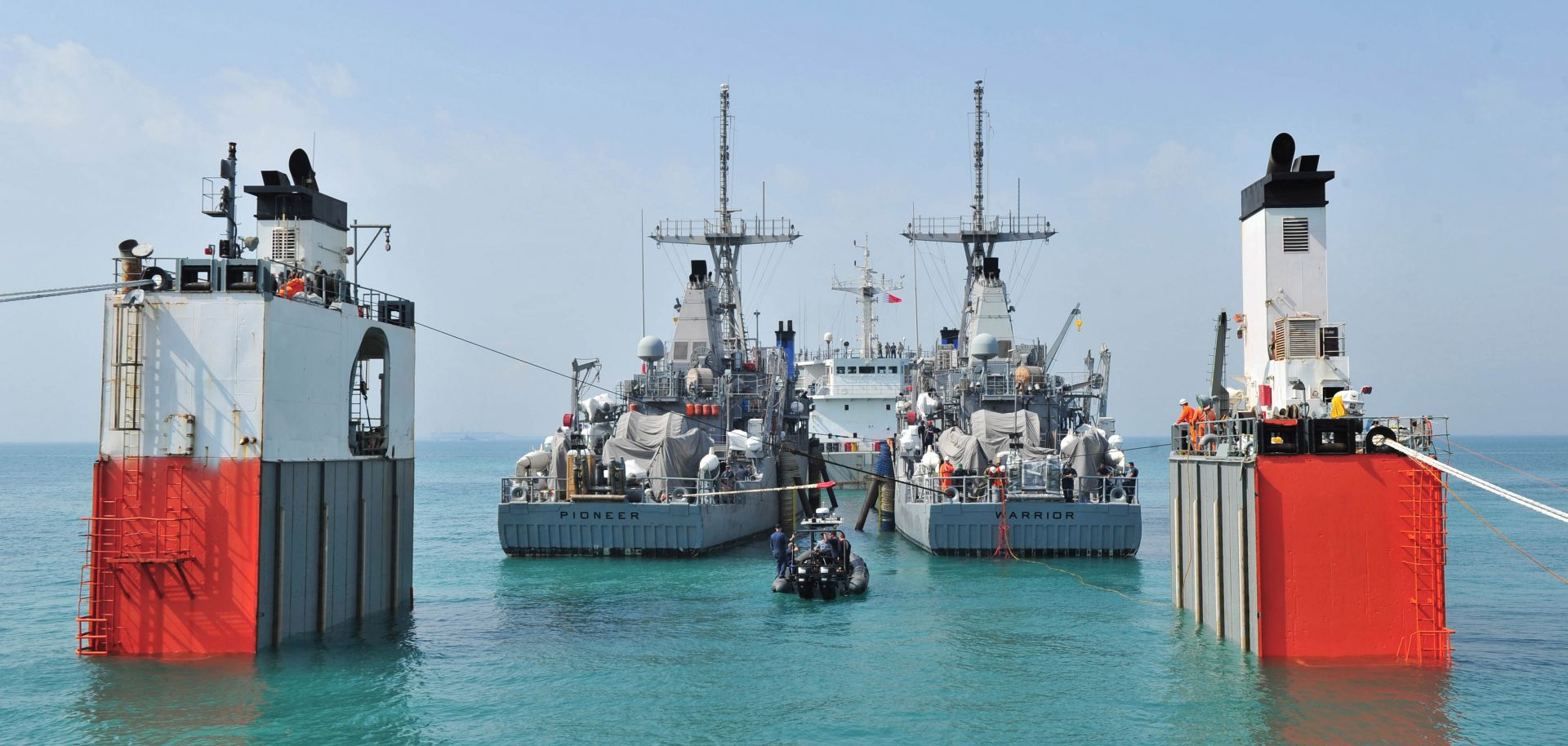 In calm turquoise waters two mine countermeasure ships USS Pioneer and USS Warrior prepare for transport.