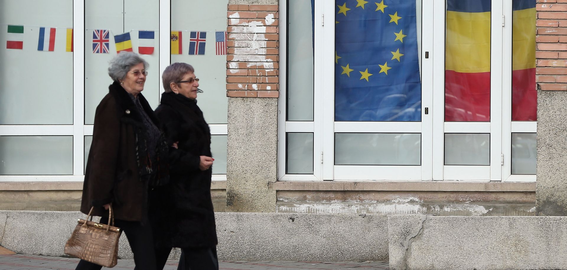 Two elderly women walk past a storefront displaying the European Union and Romanian flags.