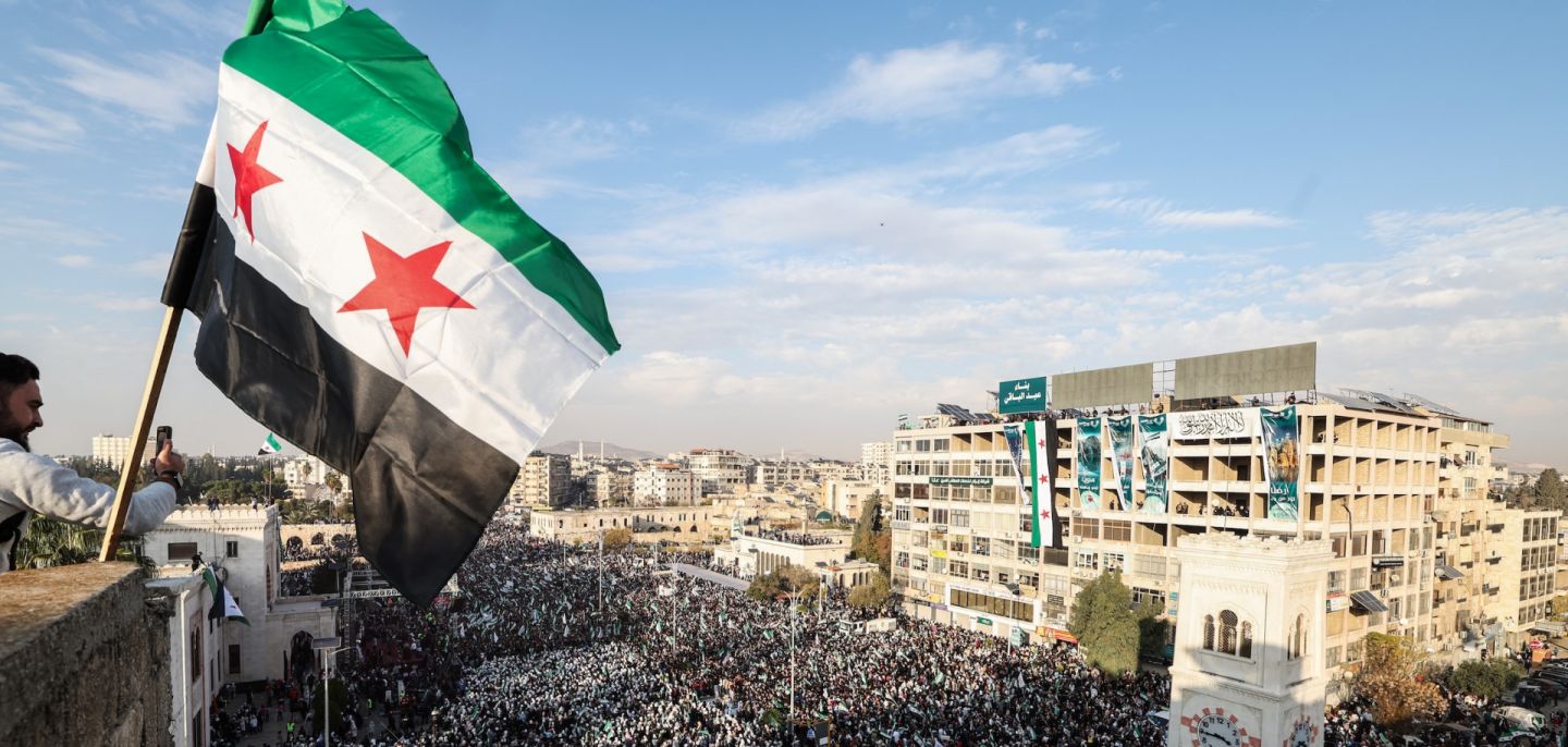 A Syrian flag flutters above crowds gathering in central Hama, Syria, during celebrations on Dec. 5, 2025, marking one year since the fall of former President Bashar al Assad's regime.