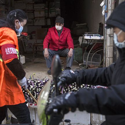 A customer examines a produce vendor's wares at a market in Wuhan, China. The economic effects of the city's quarantine on the country's larger economy will grow more pronounced the longer the lockdown stays in place. This photo shows a masked vendor and customers of his wares in an alley in Wuhan, China, on January 31, 2020.