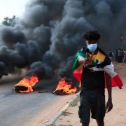 A protester draped with Sudan’s national flag stands in front of burning tires during a demonstration in Khartoum on Oct. 25, 2021. A protester draped with Sudan’s national flag stands in front of burning tires during a demonstration in Khartoum on Oct. 25, 2021.